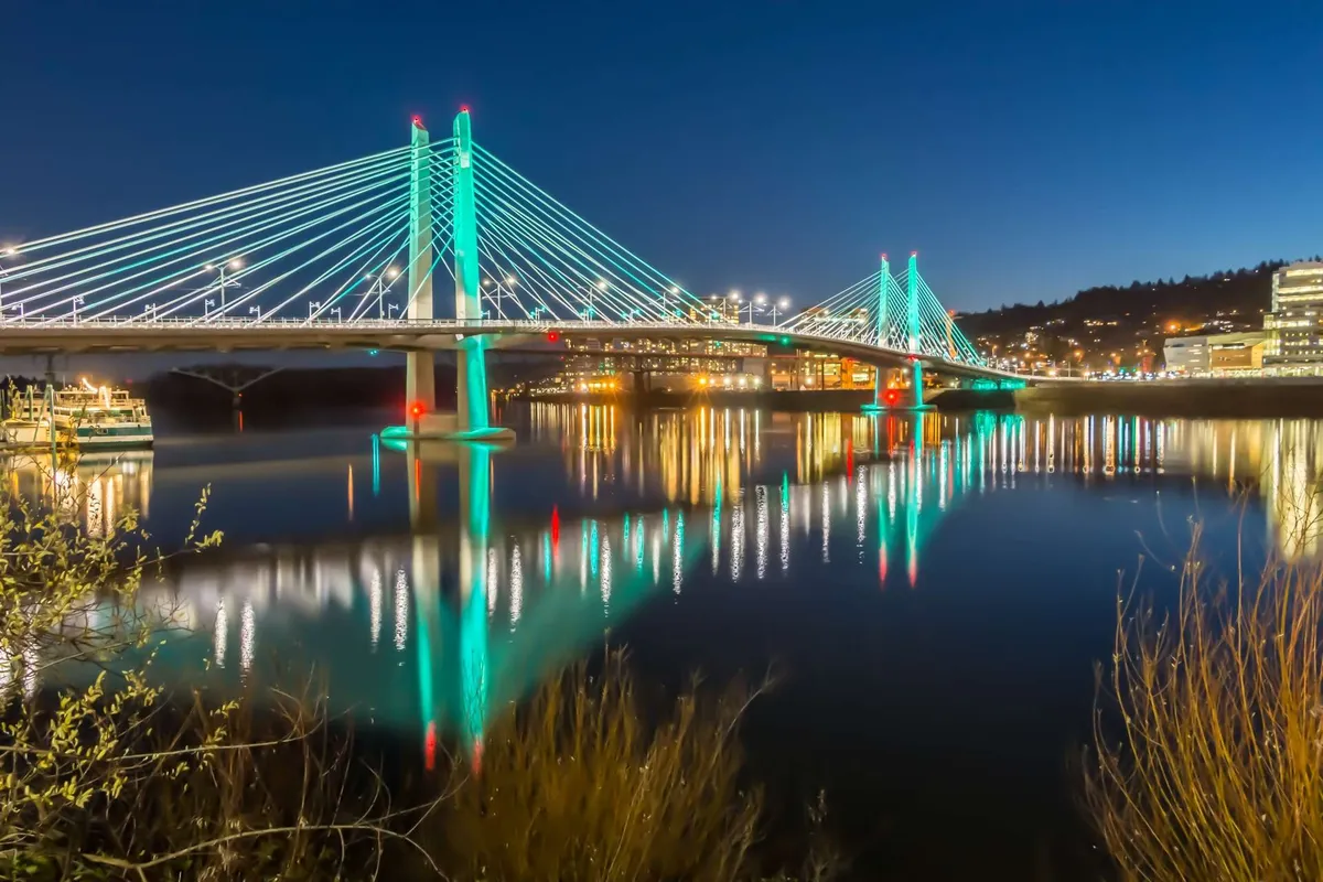 A striking evening scene featuring a bridge with illuminated cables, the city skyline reflected elegantly in the water. The vibrant yet tranquil setting captures the spirit of riverside dining, nighttime strolls, and the dynamic life along the urban waterfront.