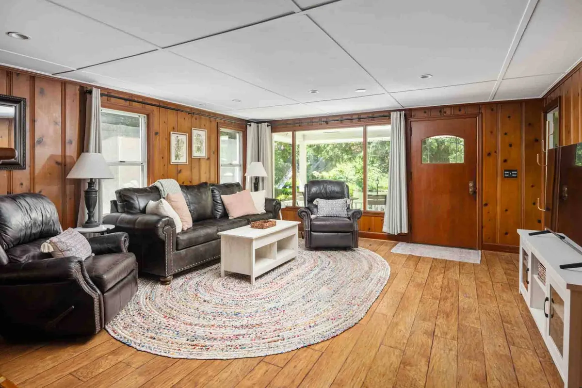 A bright, airy living area with plush leather sofas, a central coffee table, a patterned area rug, and expansive windows that let in streams of natural light. The scene suggests a space perfect for unwinding, reading, or gathering for games with loved ones.