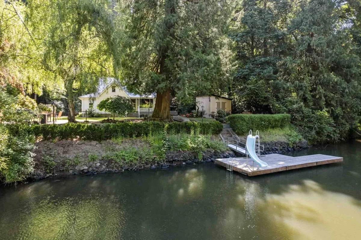 A view of the private dock extending into the river, surrounded by lush greenery and mature trees. The house is visible, and there is a swimming slide attached to the dock, with calm water inviting for swimming, boating, or fishing. The setting is tranquil, with hints of sunlight dappling through the trees.