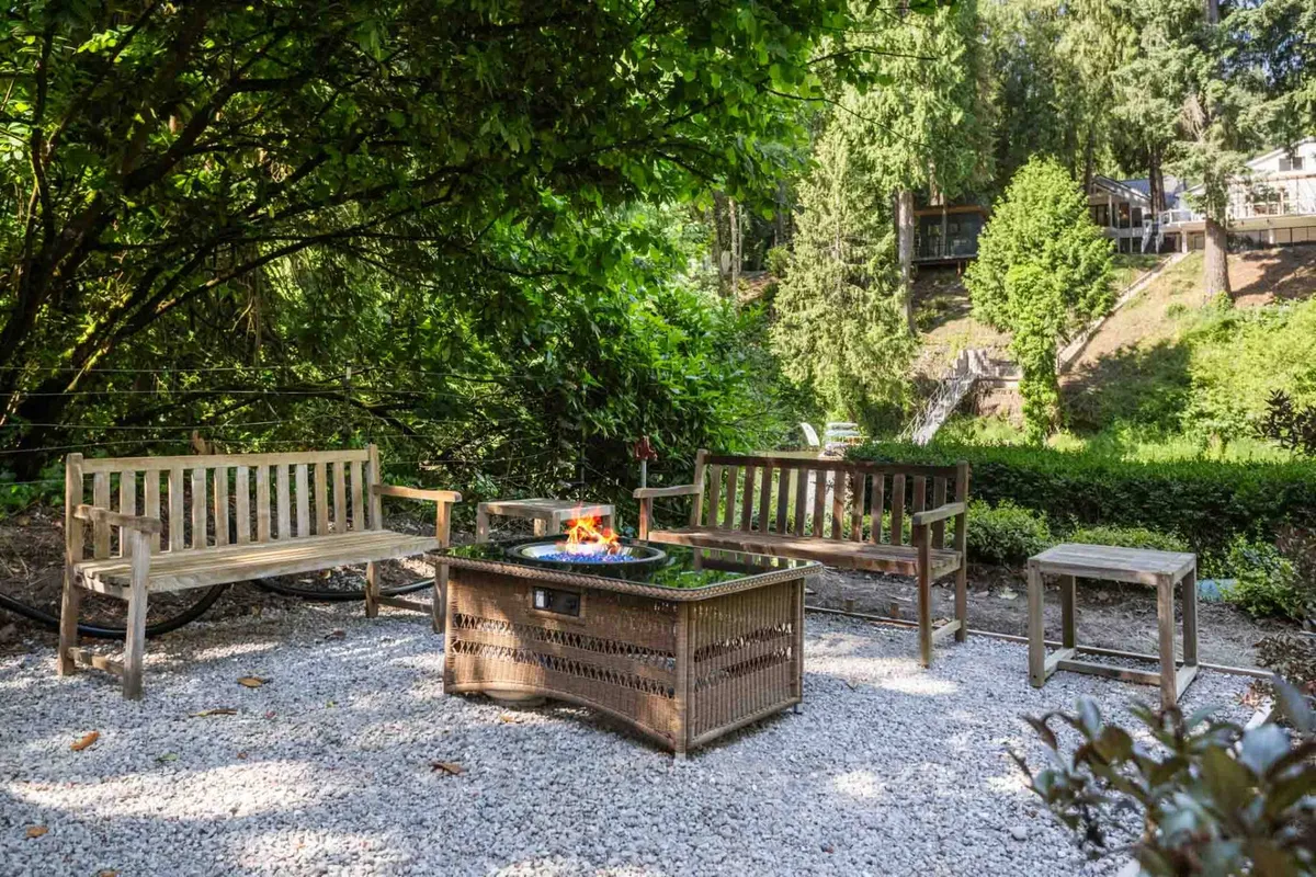 An inviting outdoor gathering spot, showcasing wooden benches arranged around a glowing fire pit on a gravel surface. The firelight creates a warm, communal ambiance, with the surrounding natural landscape visible in the background.