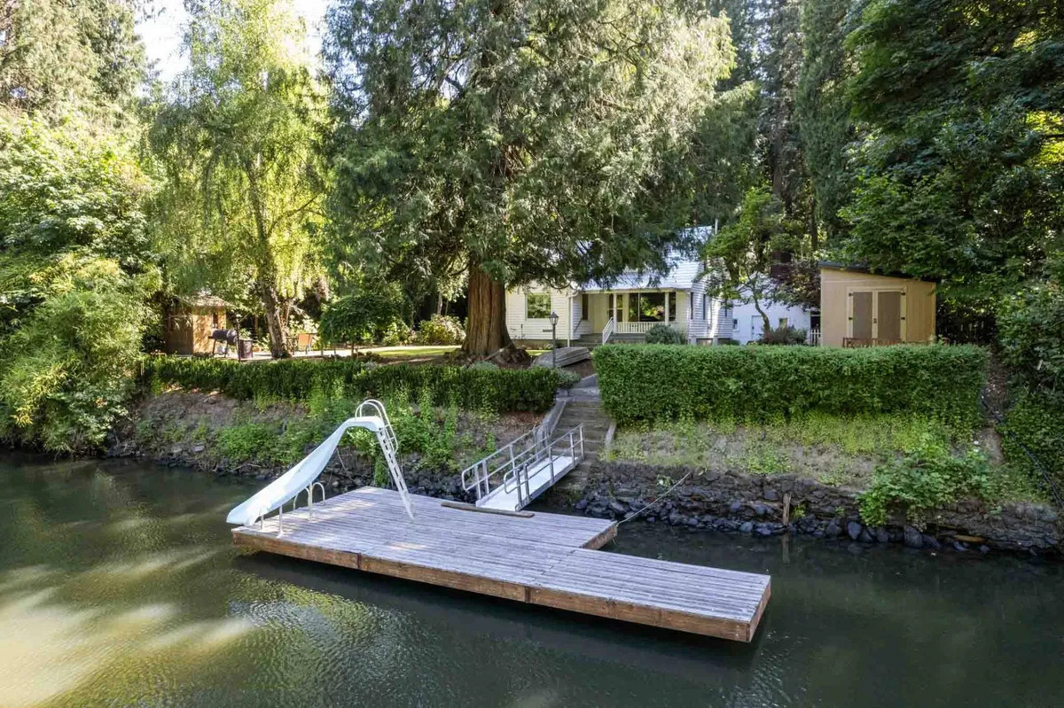 A sunny riverside scene with a large dock featuring a slide, a prominent mature tree, and the house in the background. The inviting dock is framed by lush greenery, evoking a sense of peaceful retreat and plenty of space for yoga, contemplation, or playful activities.