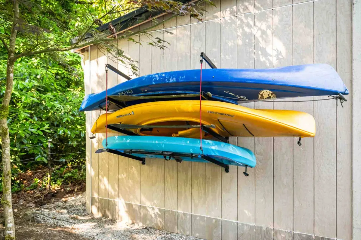 A closer look at stacked colorful kayaks resting against a wooden wall among dense greenery, suggesting easy access to water toys for guests. The scene hints at adventurous kayaking opportunities and the spirit of outdoor recreation by the water.