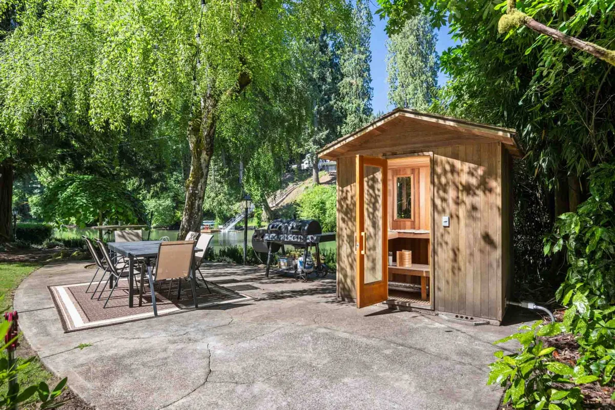 A serene outdoor area featuring a wooden sauna, an outdoor dining table with chairs, and a BBQ grill, all set beneath towering trees with lush greenery surrounding the scene. The space exudes a sense of peace and wellness, ideal for relaxation and outdoor gatherings.
