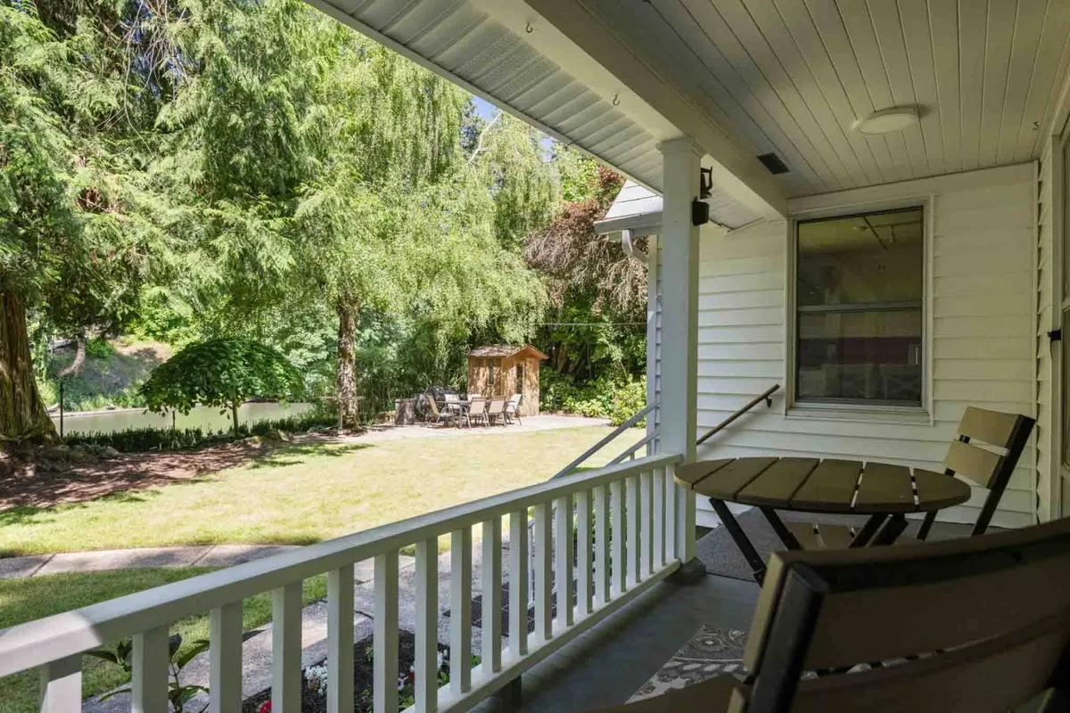 A tranquil porch scene with a sturdy railing, a table and chairs set for al fresco dining, and thriving greenery just beyond. The space suggests peaceful outdoor meals, quiet relaxation, and enjoying the passing seasons.