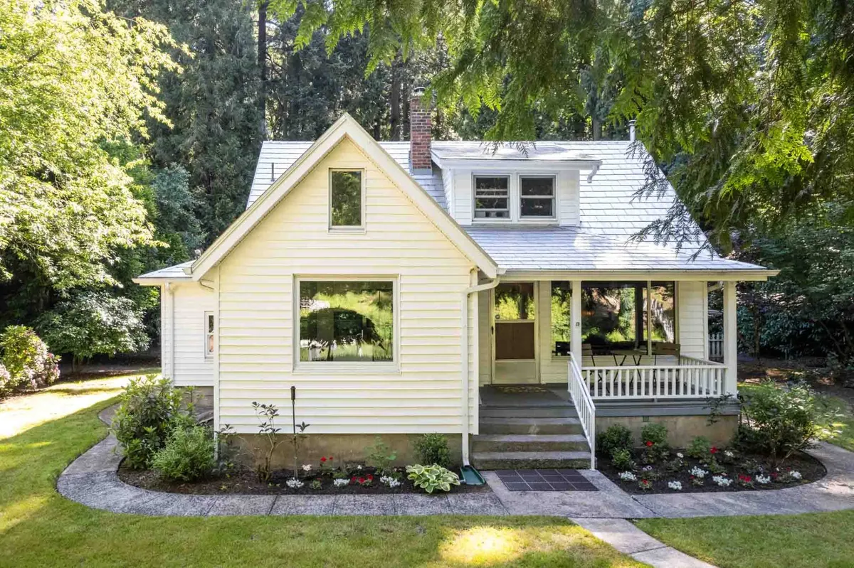 This exterior image showcases the inviting front porch of Riverwood, with large windows, well-tended flower beds, and a glimpse of the peaceful surroundings. The house exudes historic charm while appearing well-maintained and welcoming, suggesting a blend of tradition and comfort.