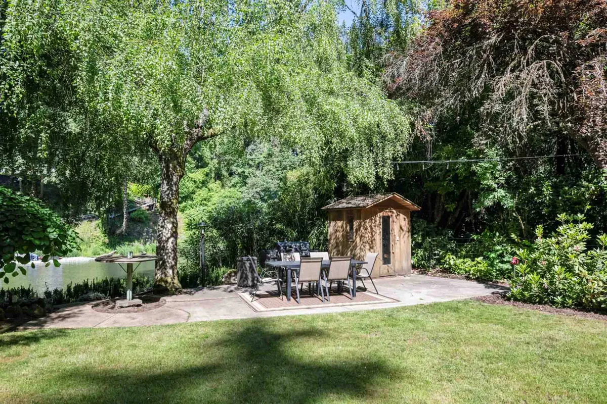 An inviting backyard scene with a dining table and chairs nestled among lush greenery and a gentle water feature. The space radiates serenity, ideal for outdoor dining, relaxing, and appreciating the quiet comforts of a riverside retreat.