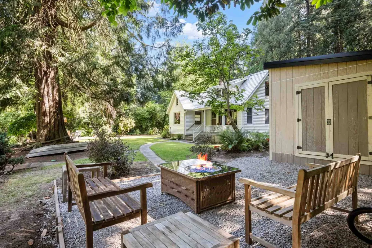 The image showcases the exterior of a charming 1930s home surrounded by lush greenery, with a cozy fire pit and wooden seating as the focal point. A winding pathway leads to the inviting house, while sunset light casts a warm glow over the outdoor gathering space. The setting suggests opportunities for outdoor dining, relaxing, and enjoying nature.