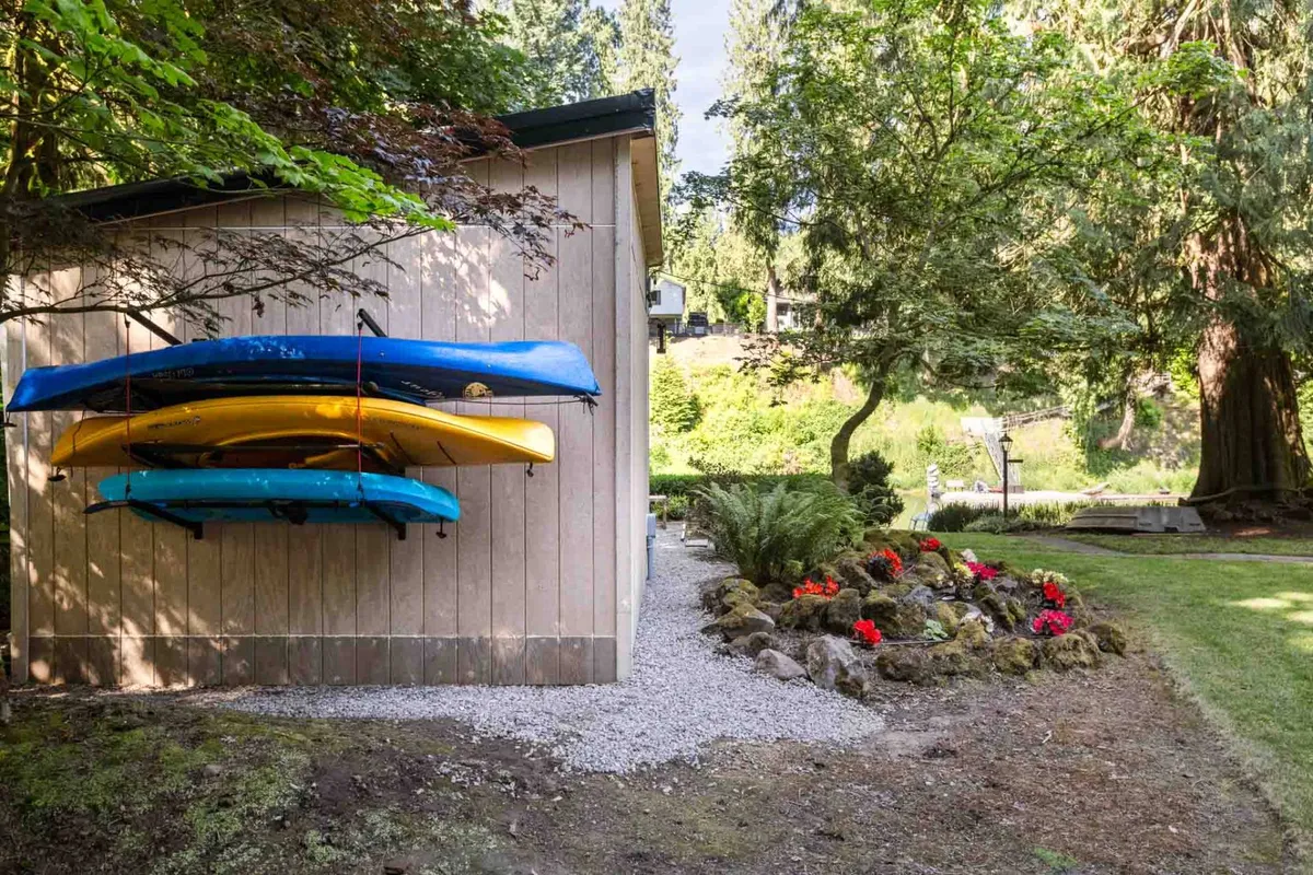 The image displays several bright kayaks stacked next to a wooden shed, bordered by a lively flower bed and lush green lawn. The surrounding landscape is vibrant and inviting, suggesting active outdoor recreation and enjoyment of the river environment.