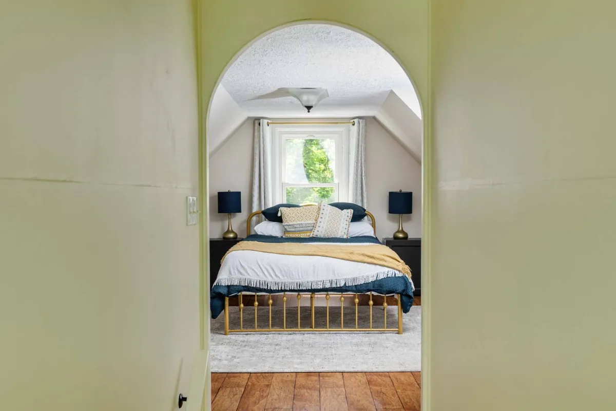 A serene bedroom with a large bed dressed in decorative pillows, bathed in natural light from a window. Nightstands with lamps flank the bed, creating a tranquil and inviting sleeping space.