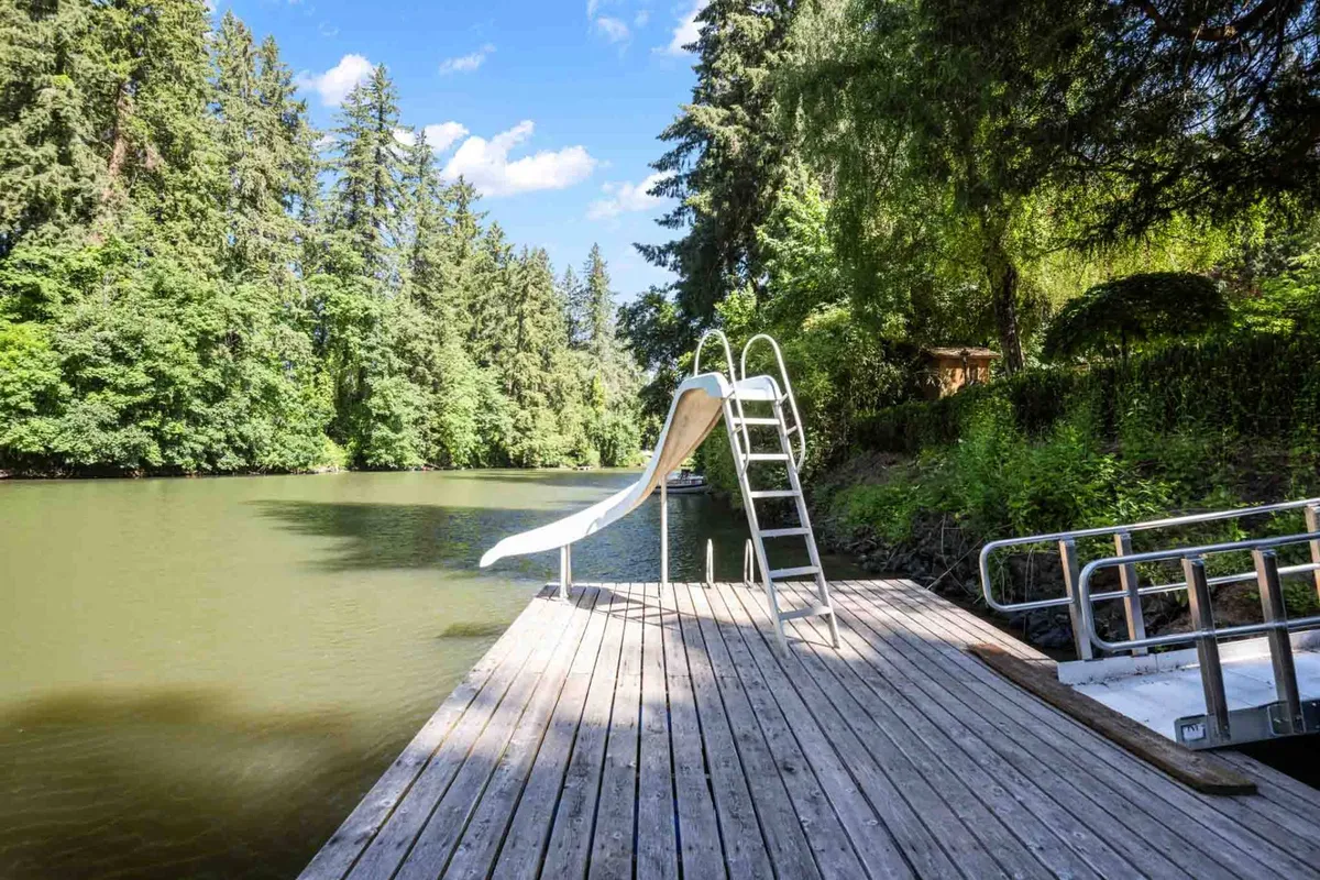 The image features a wooden dock with a water slide extending into the river, surrounded by tall trees and natural greenery. The scene radiates relaxation and outdoor enjoyment, inviting guests to experience direct river access and immersive nature activities.