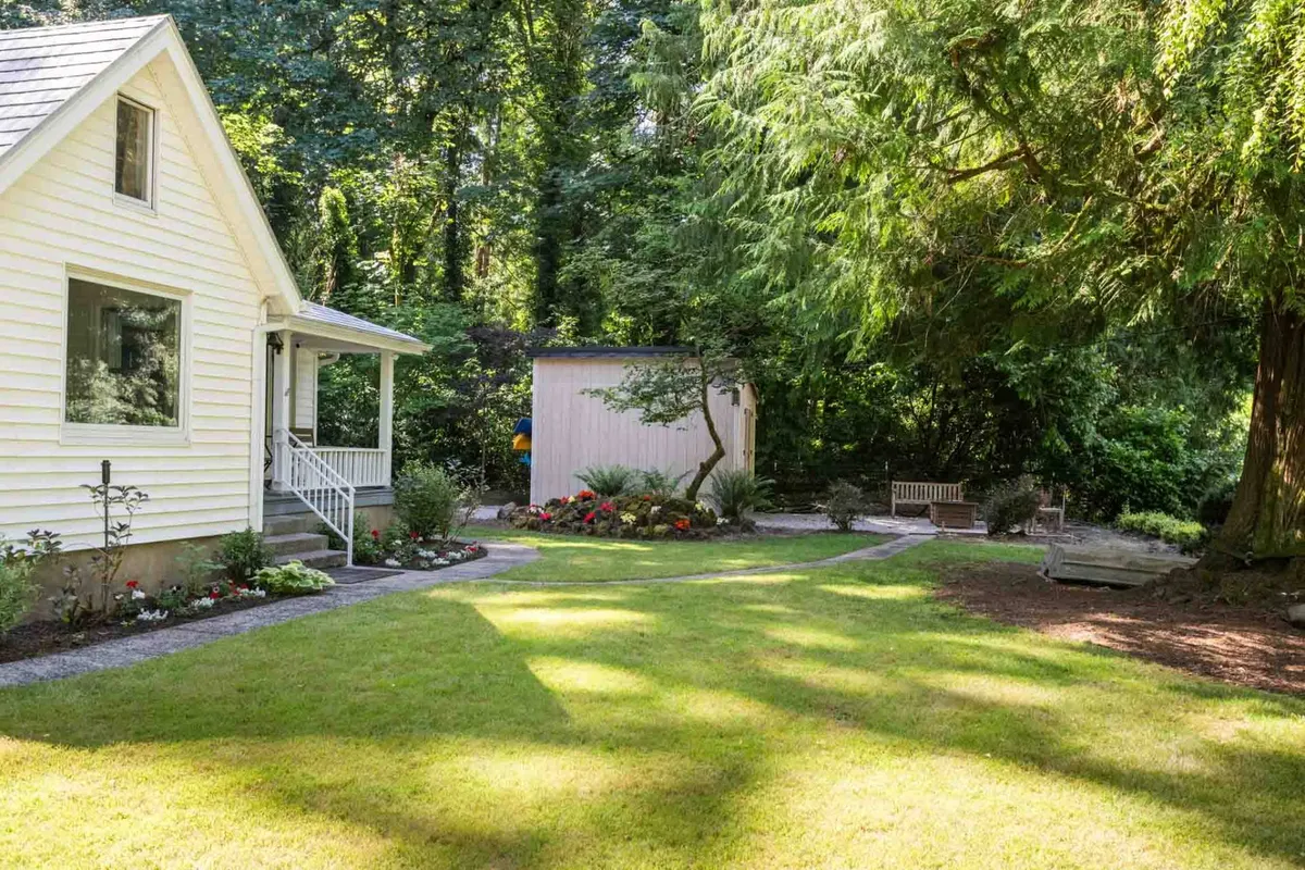 This exterior image features a white house nestled amidst a lush garden filled with colorful flowers. A wooden shed and a bench provide inviting spots within the garden, and the overall scene feels peaceful and welcoming. The open lawn and unobstructed paths suggest ample space for pets to roam and play under supervision.