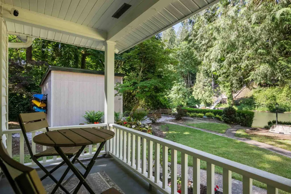 This photo shows a porch with comfortable seating, surrounded by lush greenery and winding garden paths that lead towards a water body and a shed in the background. The scene exudes tranquility, with opportunities for relaxation, picnicking, fishing, and simply enjoying the fresh air.